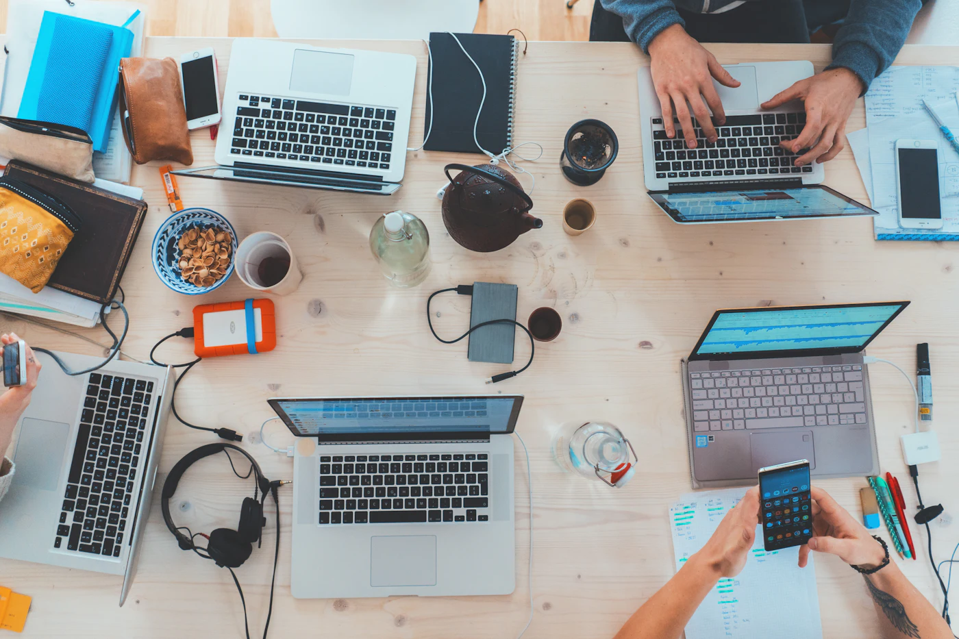 A shared desk seen from above, covered with laptops, notebooks, and phones